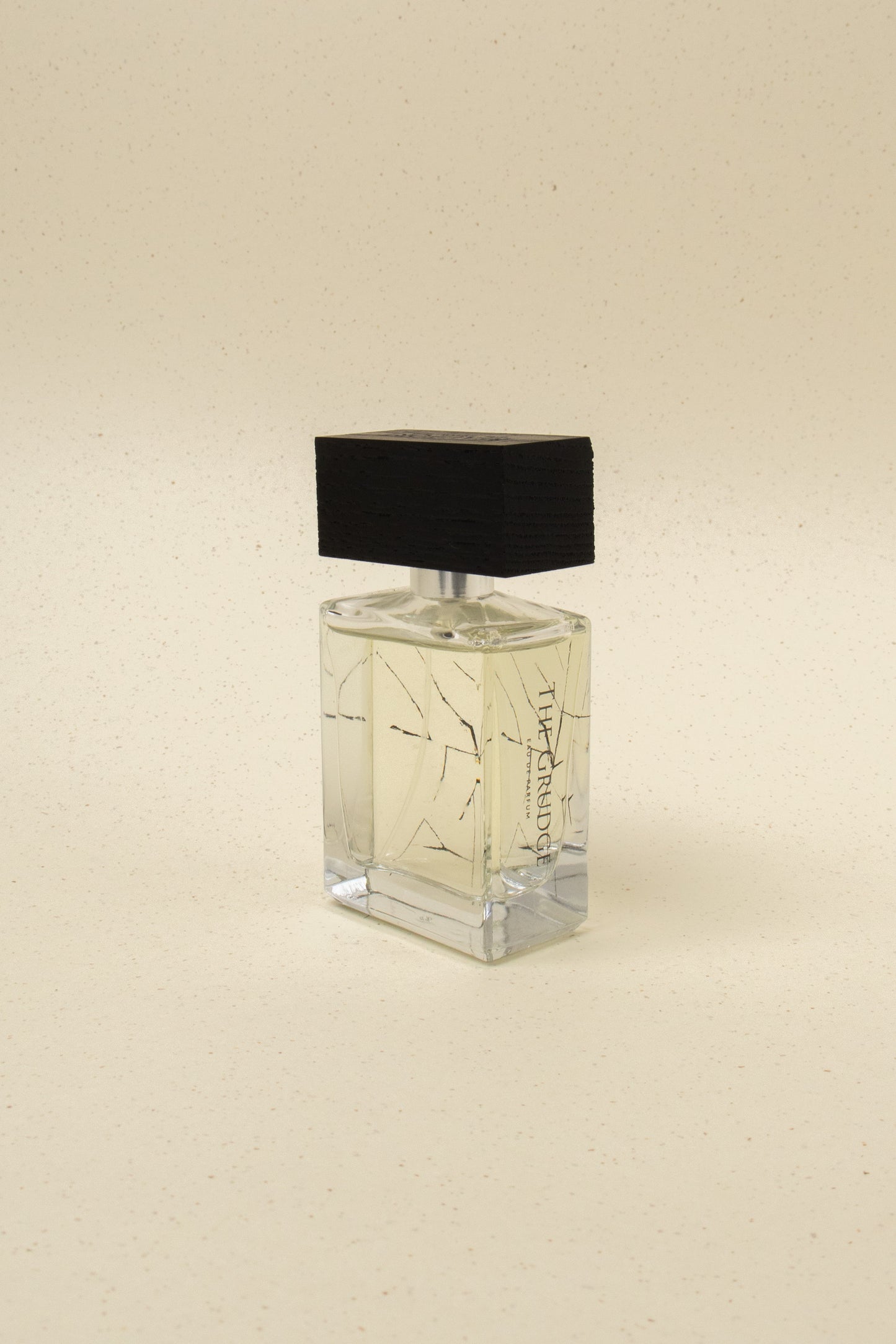 The Grudge perfume bottle with clear liquid and black wooden cap on a speckled beige background