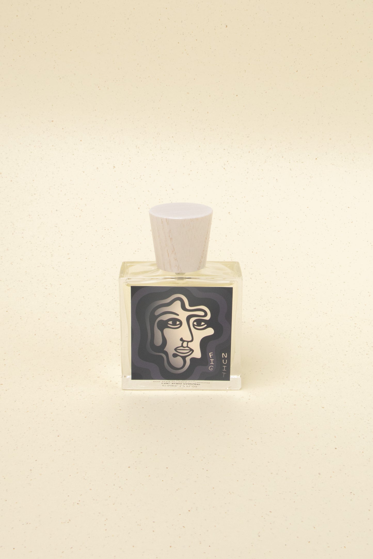 Fig Nuit perfume bottle with artistic abstract face design label and wooden cap on beige speckled background