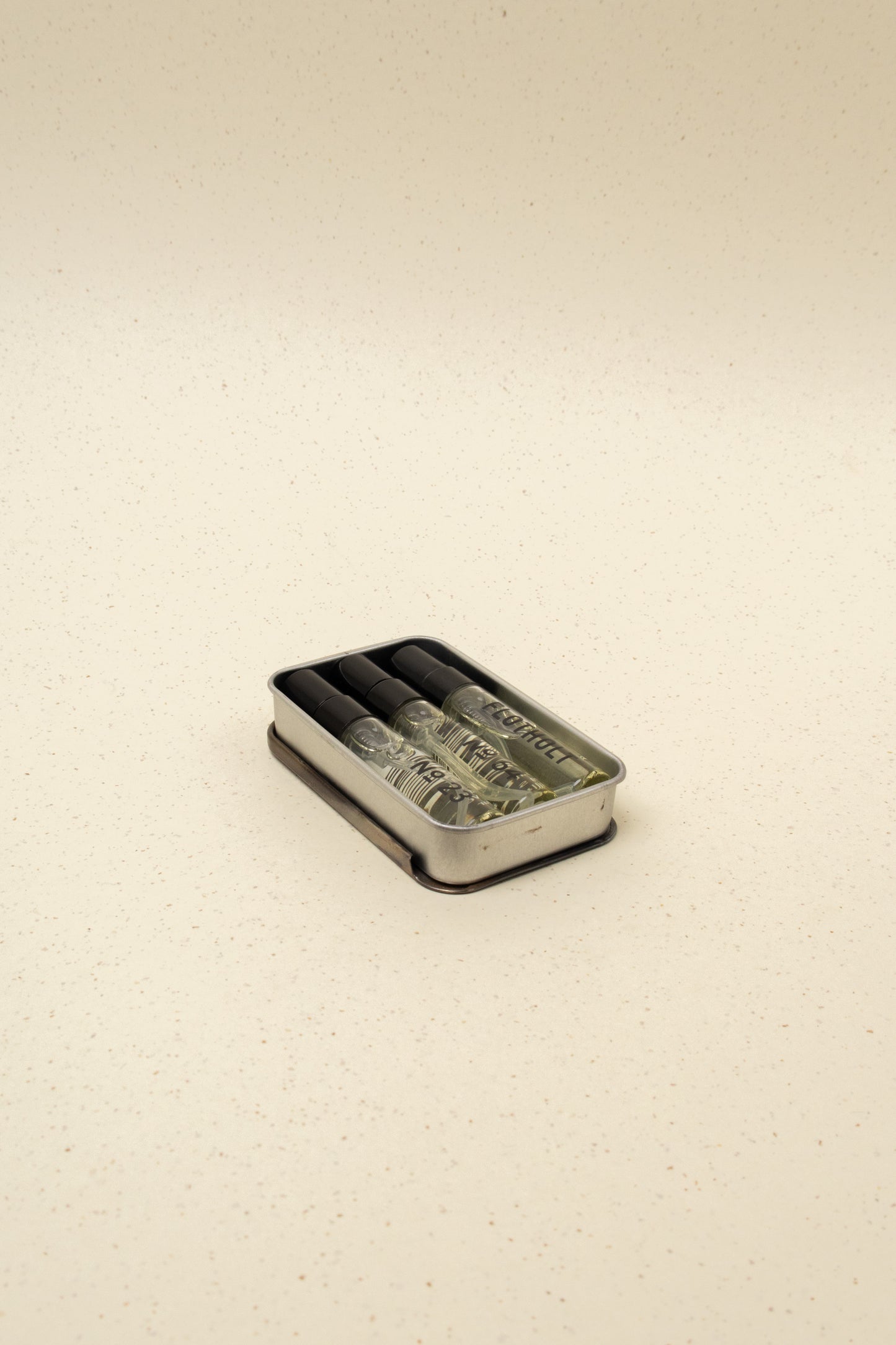 Three small glass perfume spray bottles in a rectangular metal tin on a speckled beige surface