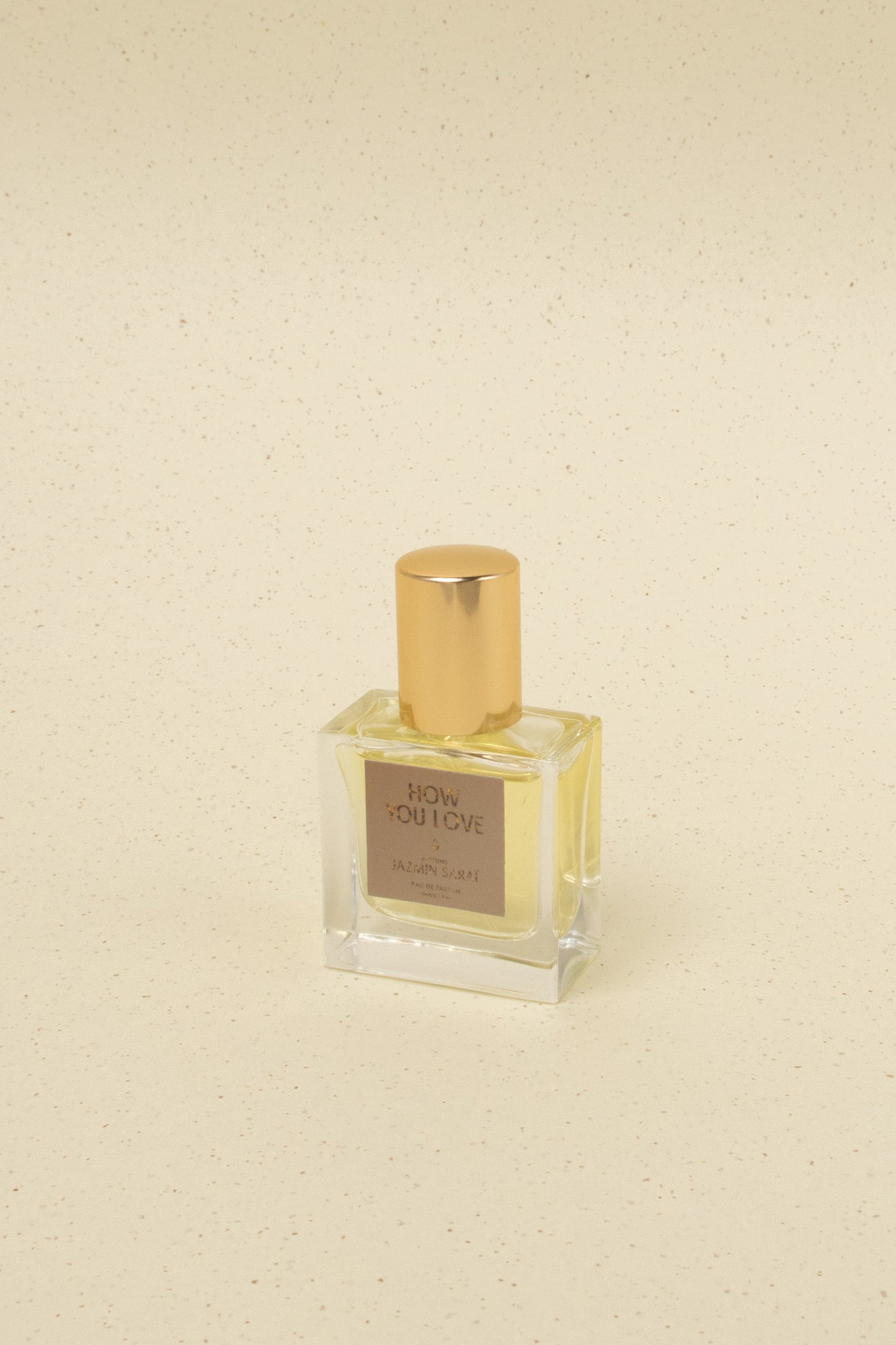 Square glass perfume bottle with gold cap and label reading How You Love by Jazmin Sarai on beige speckled background