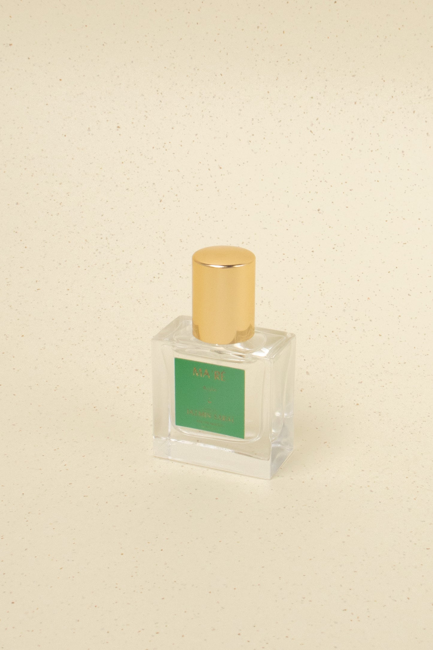 Square glass perfume bottle with gold cap and green label on beige speckled background