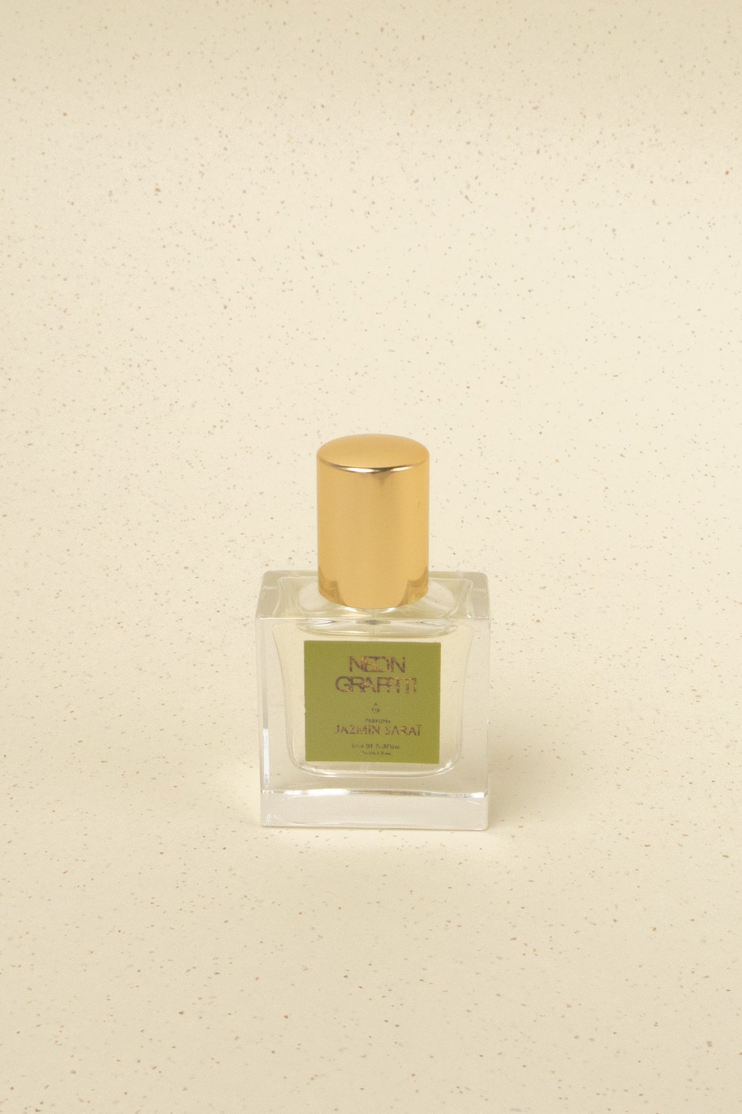 Jazmin Sarai Neon Graffiti niche perfume bottle with gold cap on speckled beige surface