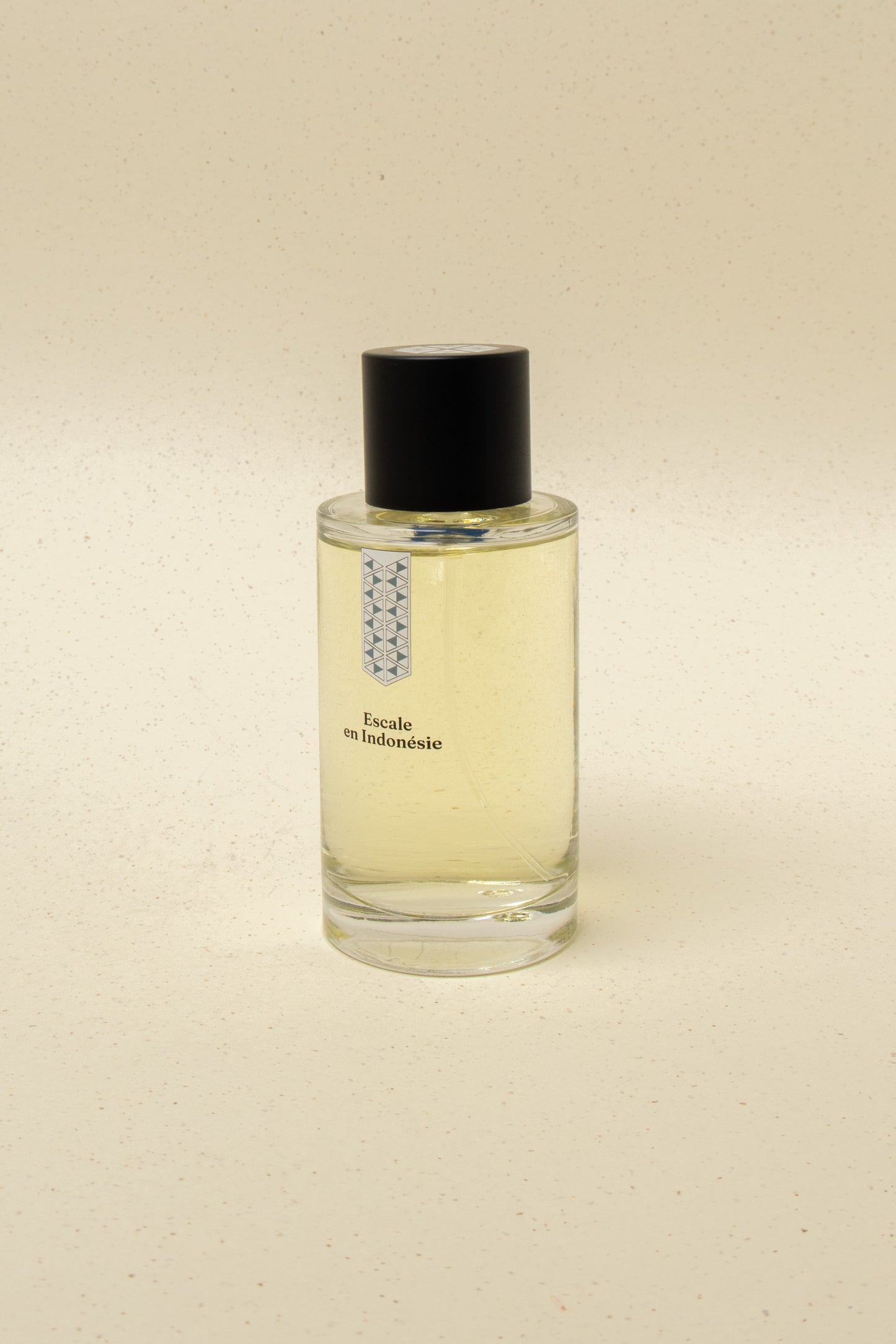 Clear glass perfume bottle with black cap labeled Escale en Indonésie on beige speckled background