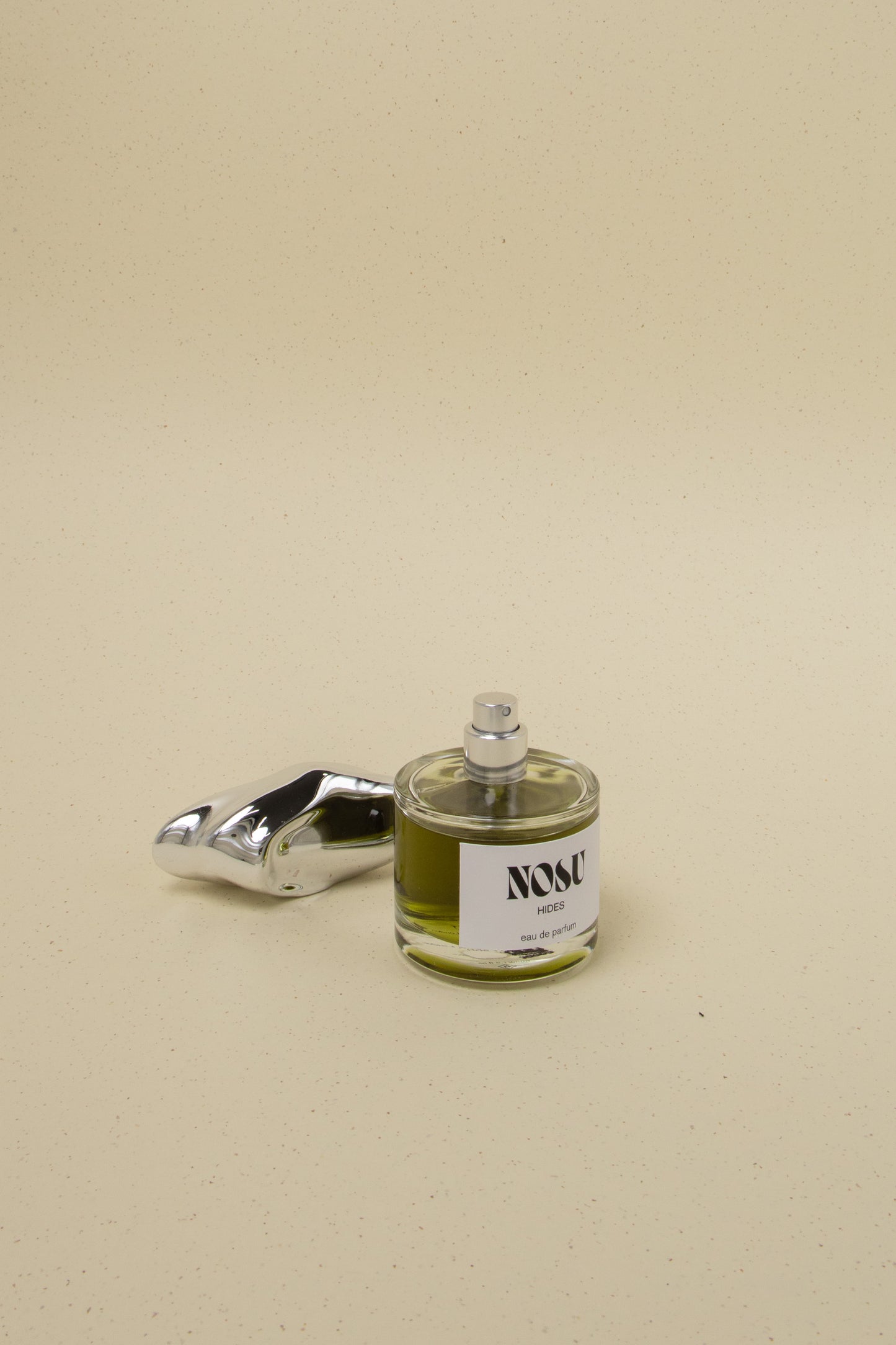 Nosu Hides eau de parfum bottle with silver sculptural cap on beige speckled background