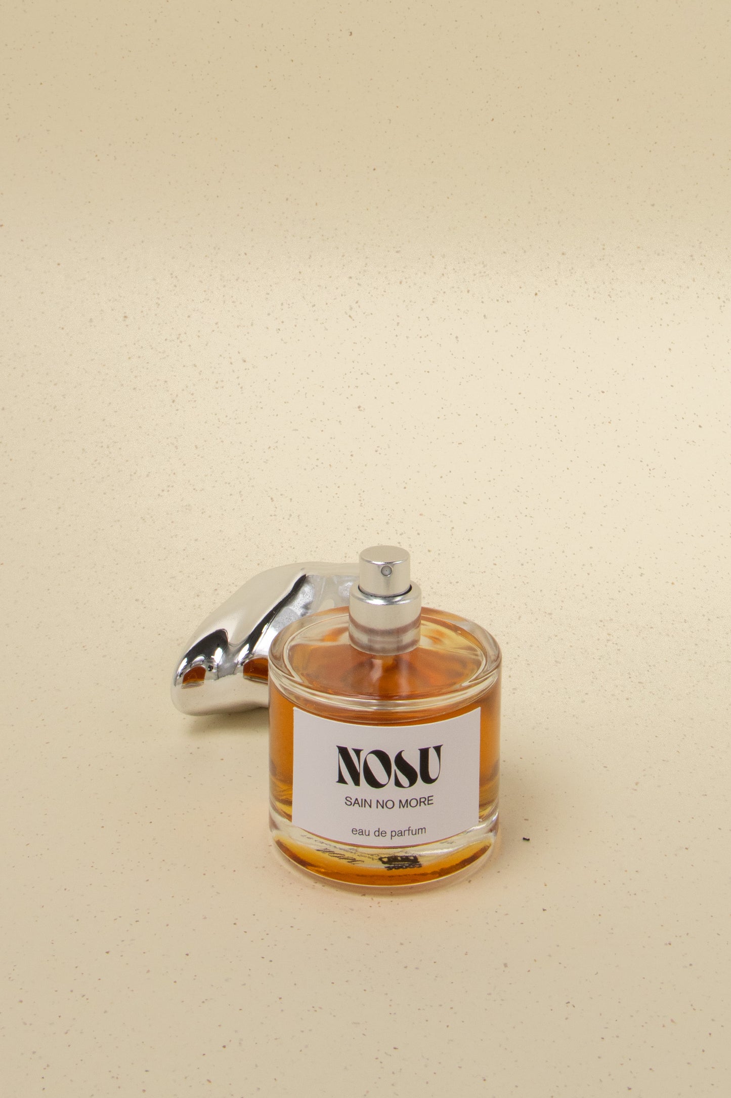 NOSU Sain No More eau de parfum bottle with amber liquid and silver spray cap on speckled beige background